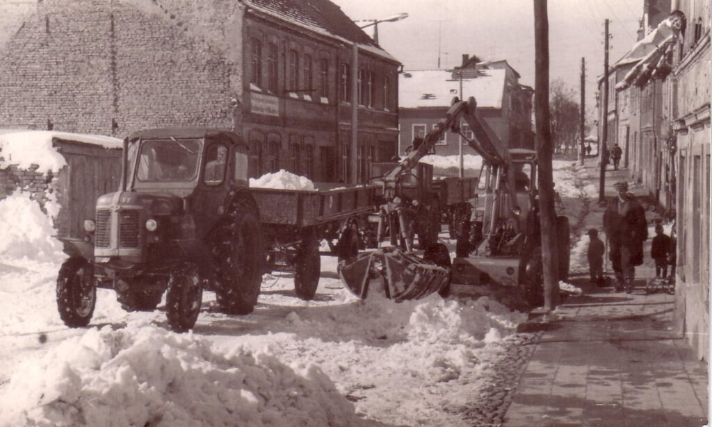 Auf dem schwarz-weiß Bild ist zu sehen, wie in der Baustraße ein Kran Schee auf einen Traktor mit Anhänger lädt. Am Bildrand sind die Schneemassen zu sehen, die vom Gehweg geschoben wurden.